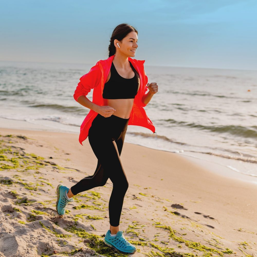 young attractive slim woman doing sport exercises in morning sunrise jogging on sea beach in sports wear, healthy lifestyle, listening to music on earphones, wearing pink windbreaker jacket